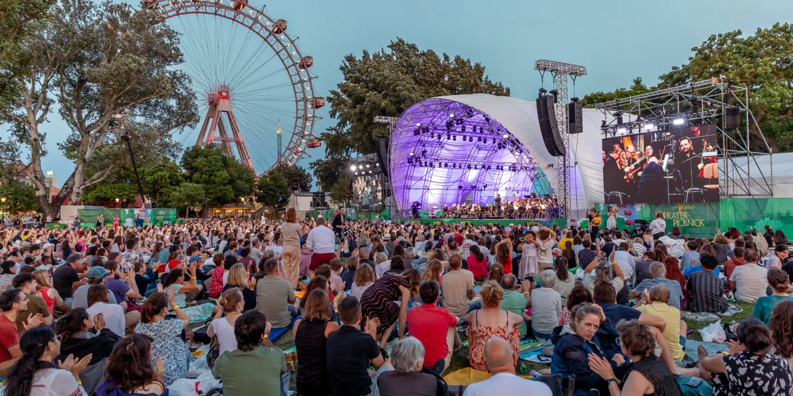Prater-Picknick der Wiener Symphoniker: Blick auf die Bühne und das Wiener Riesenrad beim Prater-Picknick der Wiener Symphoniker 2025. Auf der Kaiserwiese sitzen viele Menschen auf der Wiese und hören zu.