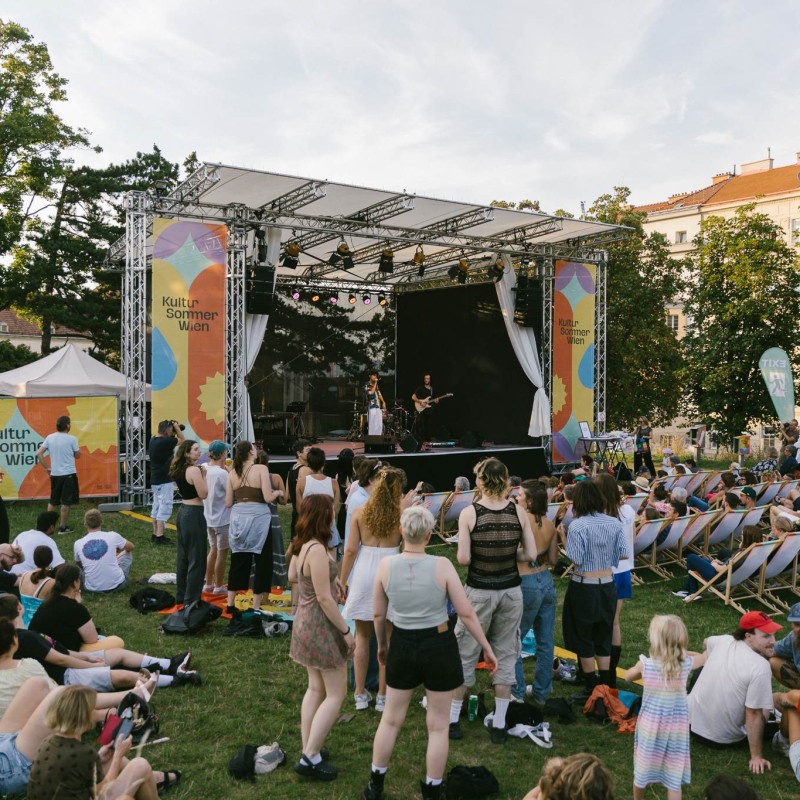: Blick auf die Bühne am Wasserturm. Das Publikum steht vor der Bühne bzw. sitzt in Liegestühlen. Auf der Bühne sind Musikerinnen zu sehen.