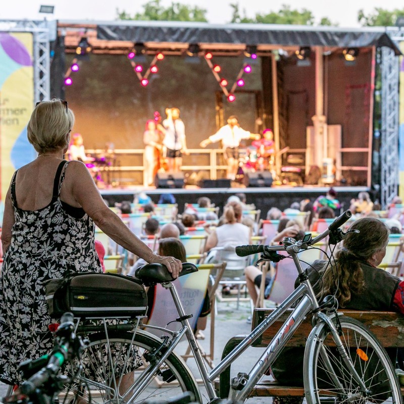 Tom Neuwrith & Martin Zerza: Blick auf die Bühne am Schrödingerplatz: Eine Dame steht mit Fahrad im Vordergrund. Das Publikum sitzt dahinter auf Liegestühlen. Auf der Bühne stehen die Musiker Tom Neuwirth und Martin Zerza mit Band.