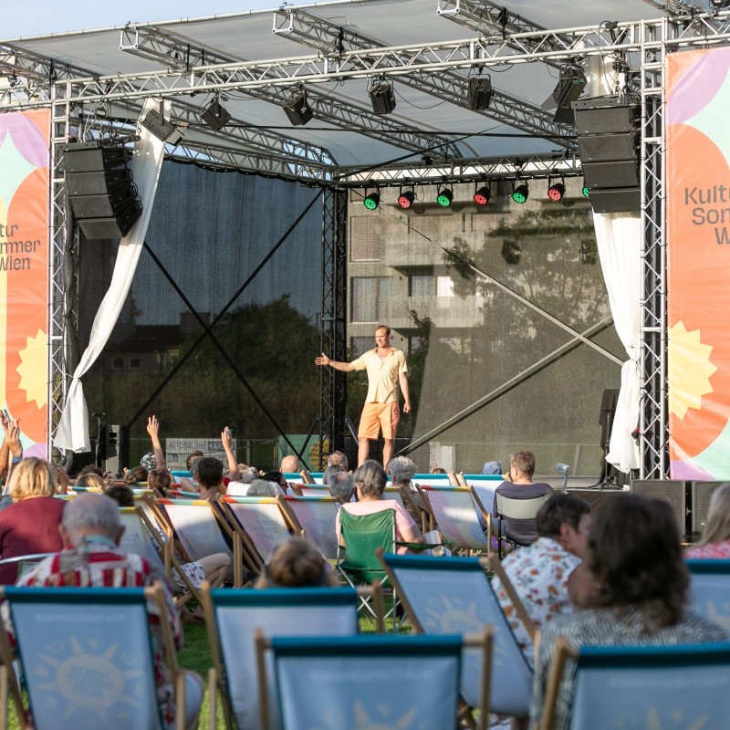 Lukas Wieser: Blick auf die Kutlursommer Bühne. Das Publikum sitzt in Liegestühlen. Lukas Wieser steht auf der Bühne.