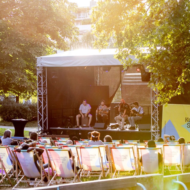 Teschek: Blick auf die Bühne im Wilhelmsdorfer Park. Das Publikum sitzt in Liegestühlen. Auf der Bühne spielt die Band Teschek ein Konzert.